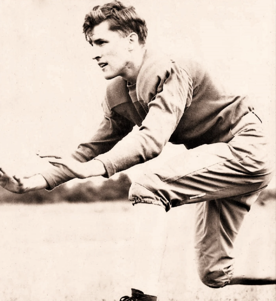 A vintage black and white image of a male athlete in mid-action pose, arms extended forward and one leg raised. He wears a light-colored, long-sleeved uniform and cleats, standing on a grassy field with a blurred background.