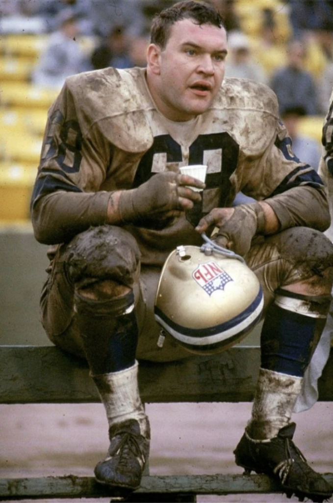 A football player in a muddy uniform sits on a bench holding a cup. His helmet, resting on his knees, has grass stains and the NFL logo. The background shows stadium seating.