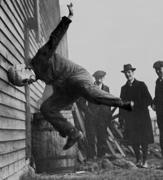 Black and white photo of a man in a mid-air flip, kicking a wall. He wears a hat and suit. Three men in hats and coats watch from the background. A barrel and wooden fence are beside the wall.