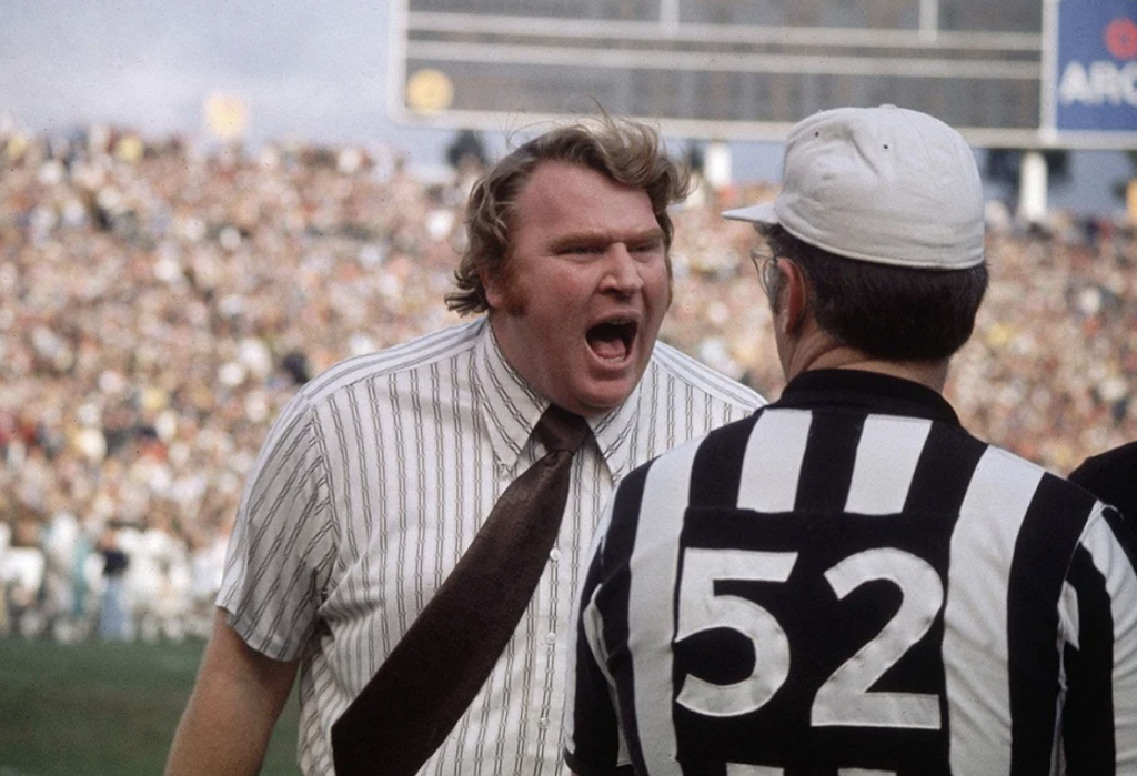 A football coach wearing a striped shirt is animatedly speaking to a referee in a striped uniform with "52" on the back. The scene takes place on a crowded sports field, with the stands filled with spectators.