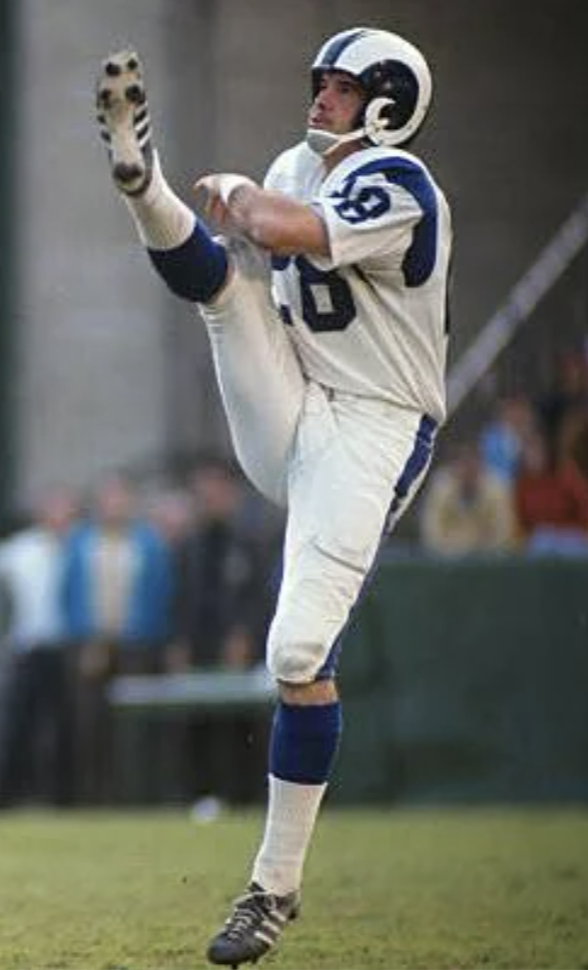 A football player in a white and blue uniform kicks a football high into the air. He is wearing a helmet with a ram horn design. Spectators in blurred focus are visible in the background.