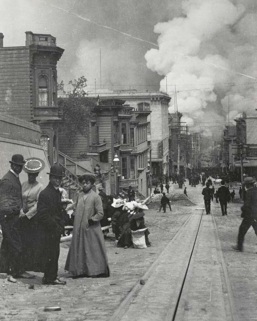 A historic black and white photo shows a busy city street with men and women in early 20th-century attire. Smoke rises in the distance, and people are gathered along the sidewalks and streetcar tracks. Buildings line both sides of the street.