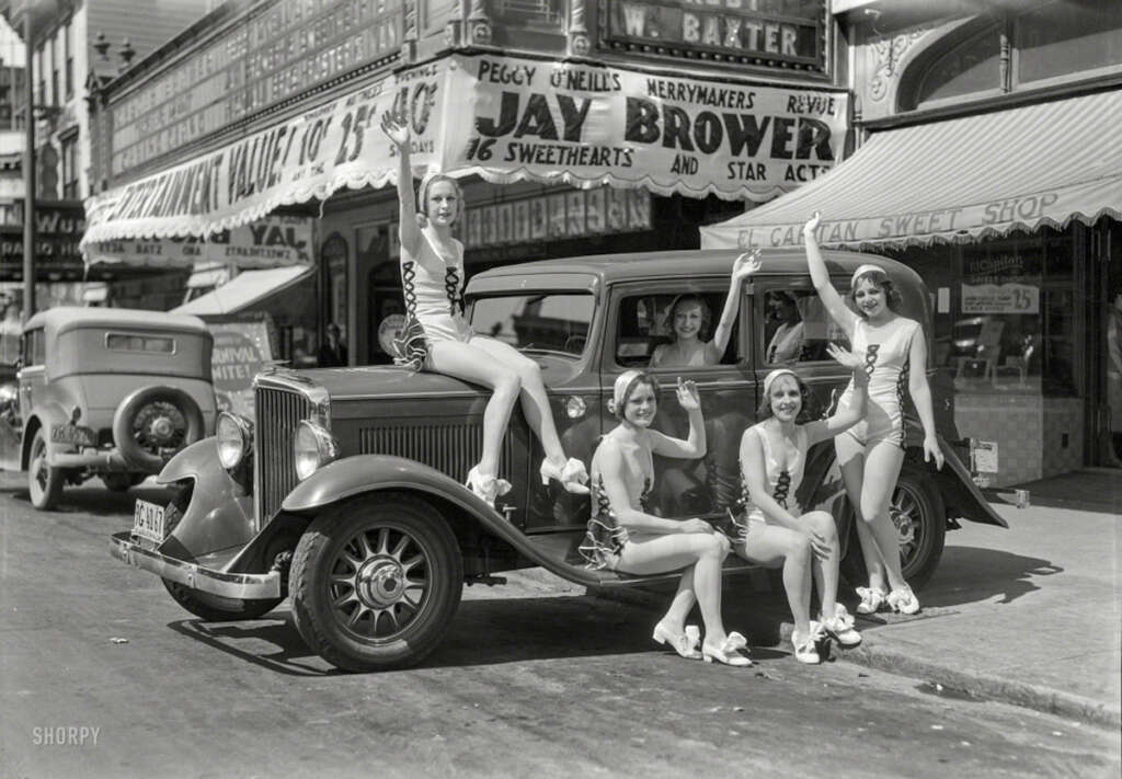 Five women in matching outfits pose on and around a vintage car parked on a street lined with shops and signs. The background features a theater marquee and storefronts with vintage advertisements.