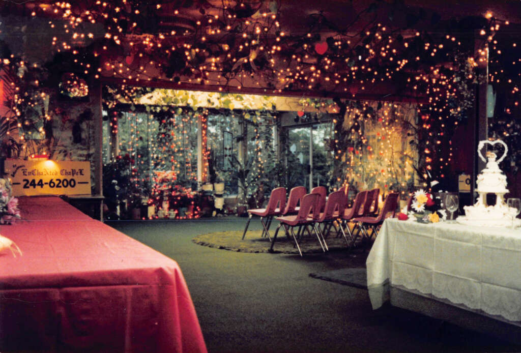 A dimly lit room decorated for a celebration with string lights and floral arrangements. Rows of red chairs face a table adorned with a multi-tiered white cake. A sign with a phone number is visible on the left side.