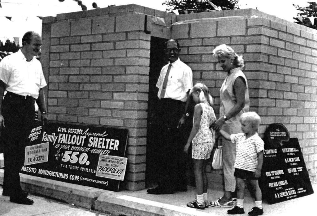 A black-and-white photo showing a family of four, two adults and two children, standing outside a brick fallout shelter. Signs advertise the shelter as a civil defense provision, pricing it at $550, by Asbesto Manufacturing Corp.