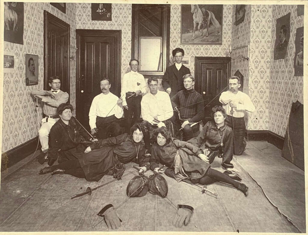 A vintage photograph depicts a group of men and women in a room with patterned wallpaper and framed pictures. They are dressed in historical fencing attire, with some holding practice swords and masks lying on the floor.