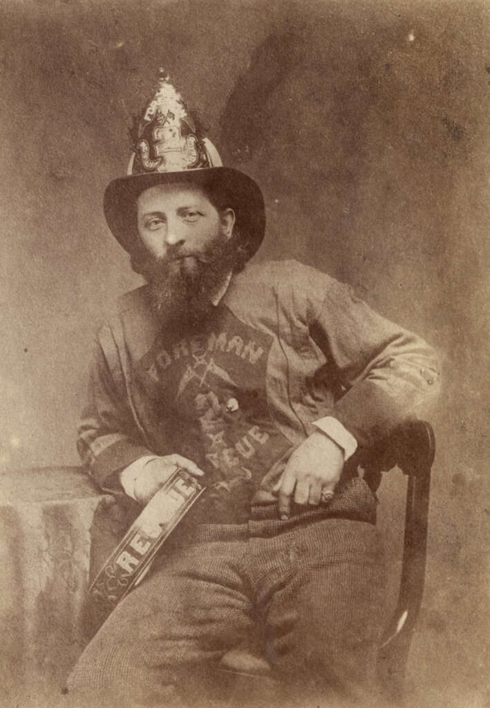 A vintage photograph of a man sitting on a chair wearing a large hat decorated with an emblem. He has a beard and is dressed in a long-sleeved shirt with "Foreman" written on it. A rolled banner with "Relief" is on his left arm.