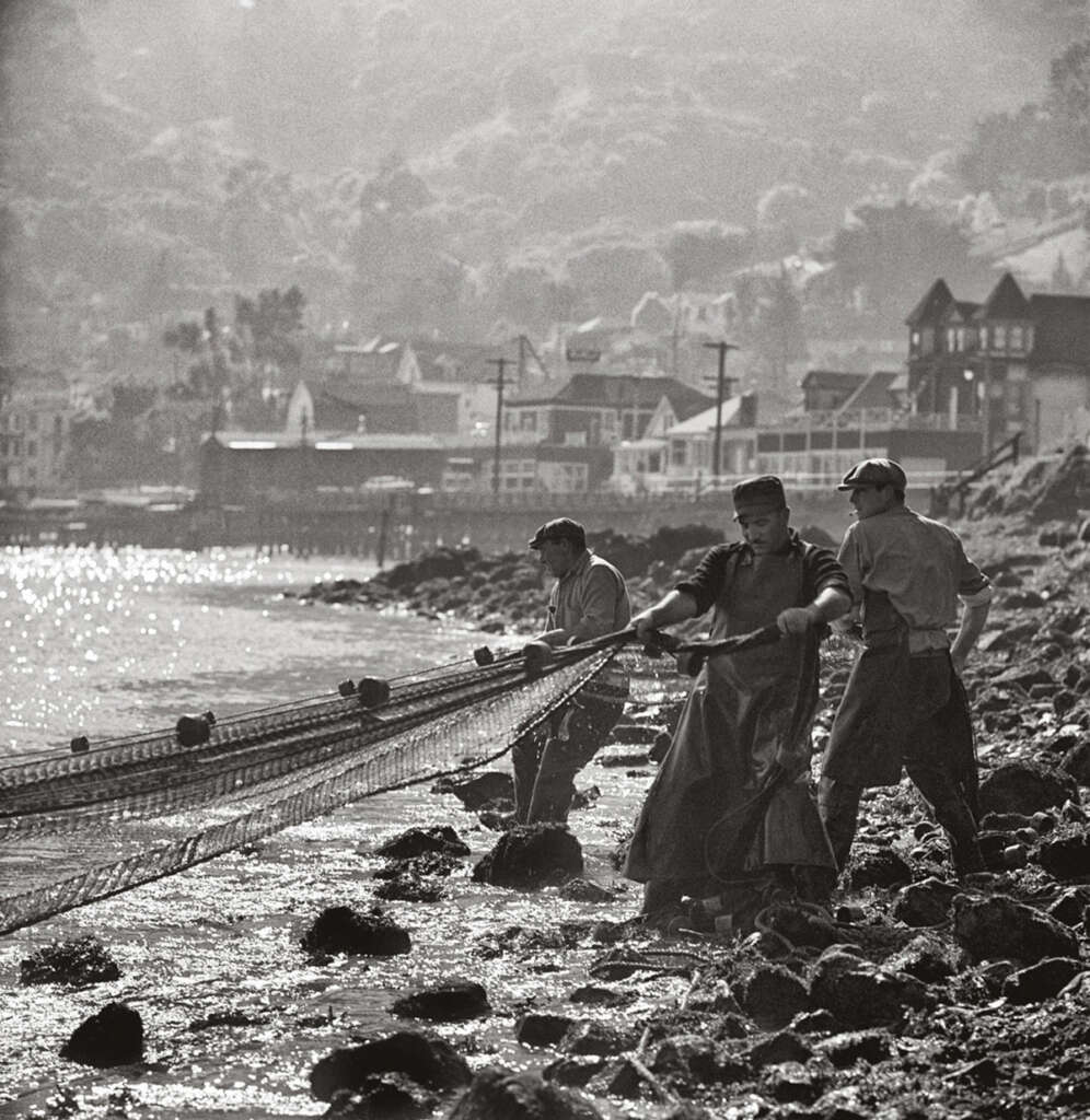 Black and white photo of three fishermen pulling a fishing net from the shore amid rocks and water. The background shows a coastal village with several buildings. The scene is lit by soft, diffused light.