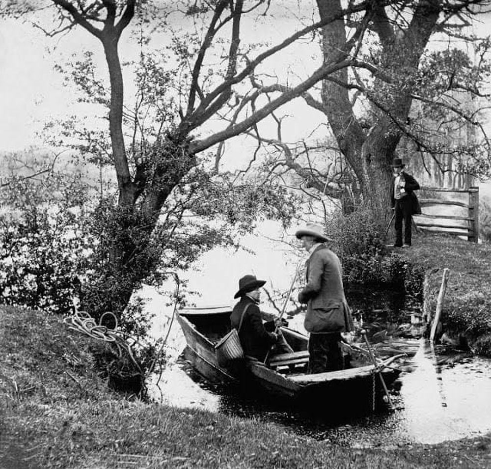 Two men wearing hats are in a small rowboat on a narrow, tree-lined waterway. A third man stands on the grassy bank nearby, observing. The scene is set in a rustic, serene outdoor environment.