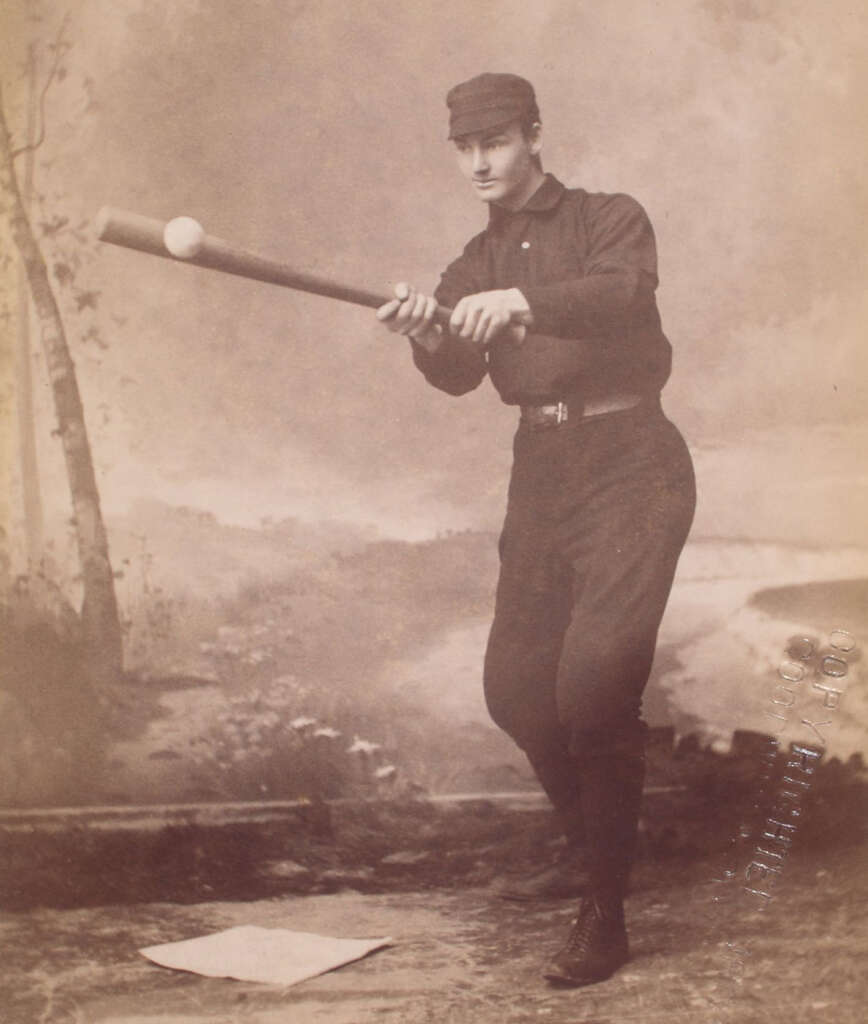 A vintage photograph of a baseball player in a dark uniform and cap, posed mid-swing with a bat, against an outdoor backdrop. The player's stance suggests readiness to hit a ball. A base is visible on the ground in front of him.