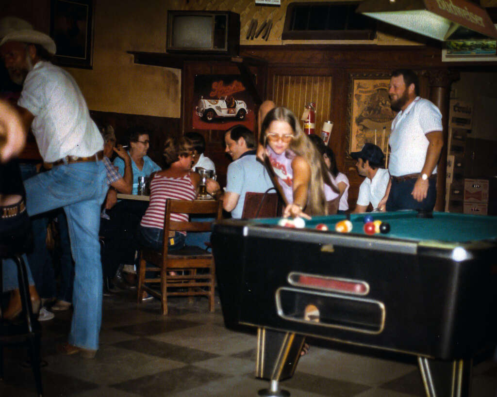 A lively bar scene with people gathered around tables and a pool table. A woman wearing sunglasses is playing pool in the foreground. Patrons in casual attire are enjoying drinks and socializing. The decor is rustic with a vintage car poster on the wall.