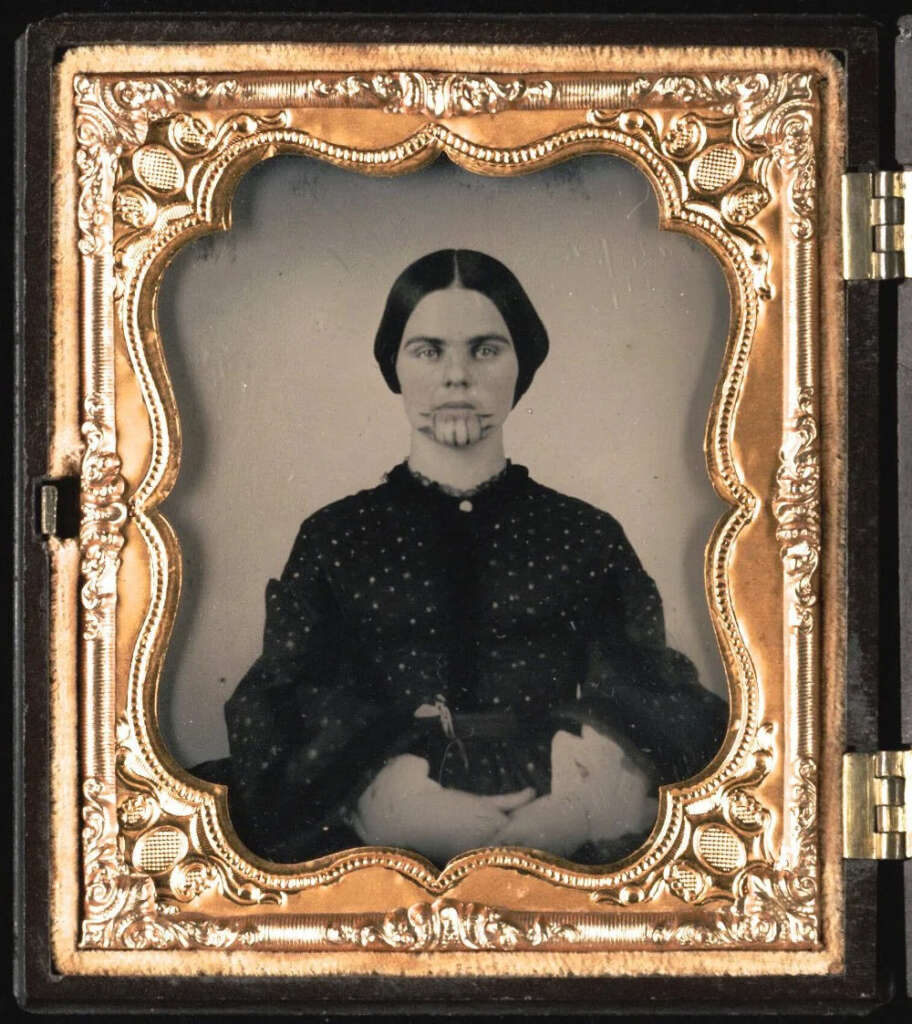 A vintage photograph of a woman with dark hair, center-parted and tied back, wearing a dark dress with white patterns. She has distinct chin tattoos and is seated with her hands crossed. The photo is in an ornate gold frame.