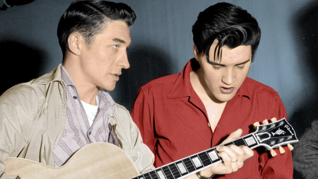 Two men are in a room; one is playing a guitar. The man on the left has short dark hair and is wearing a light-colored jacket and patterned shirt. The man on the right has dark hair and is wearing a red shirt, looking down at the guitar.