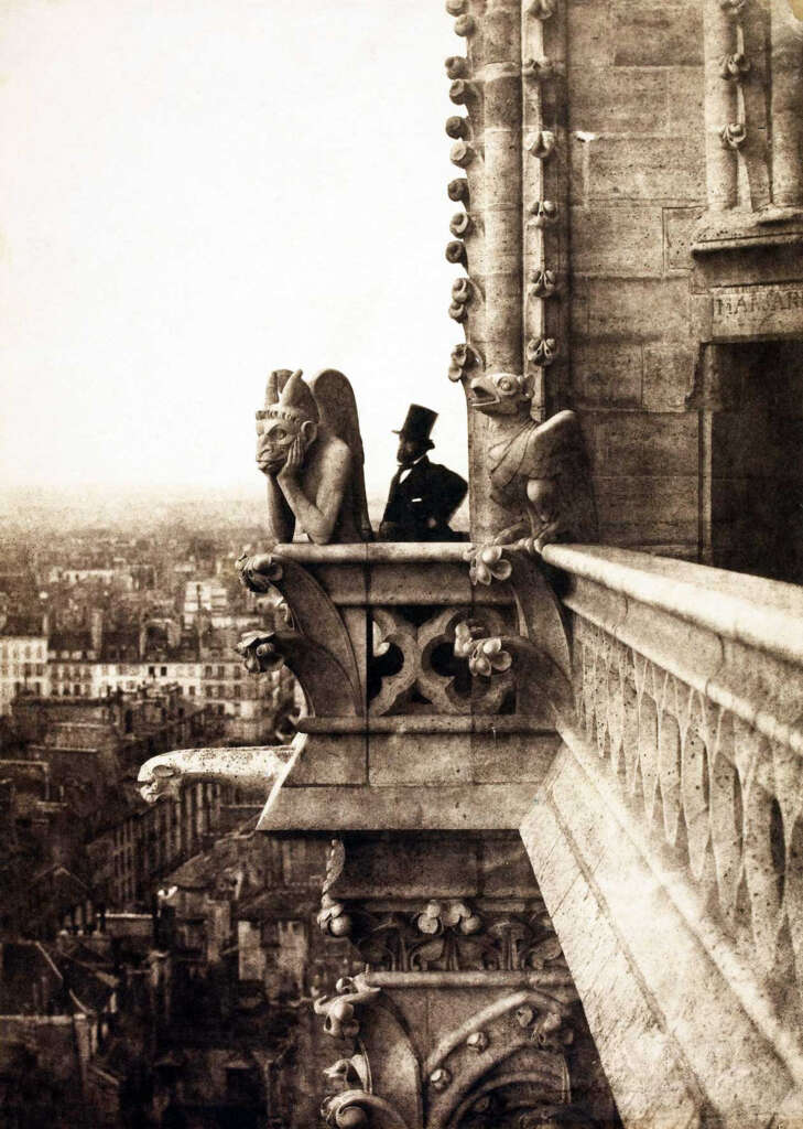 A man in a top hat stands on a balcony next to stone gargoyles overlooking a cityscape with historic buildings. The image is sepia-toned, giving it a vintage feel, and the scene suggests a high vantage point.