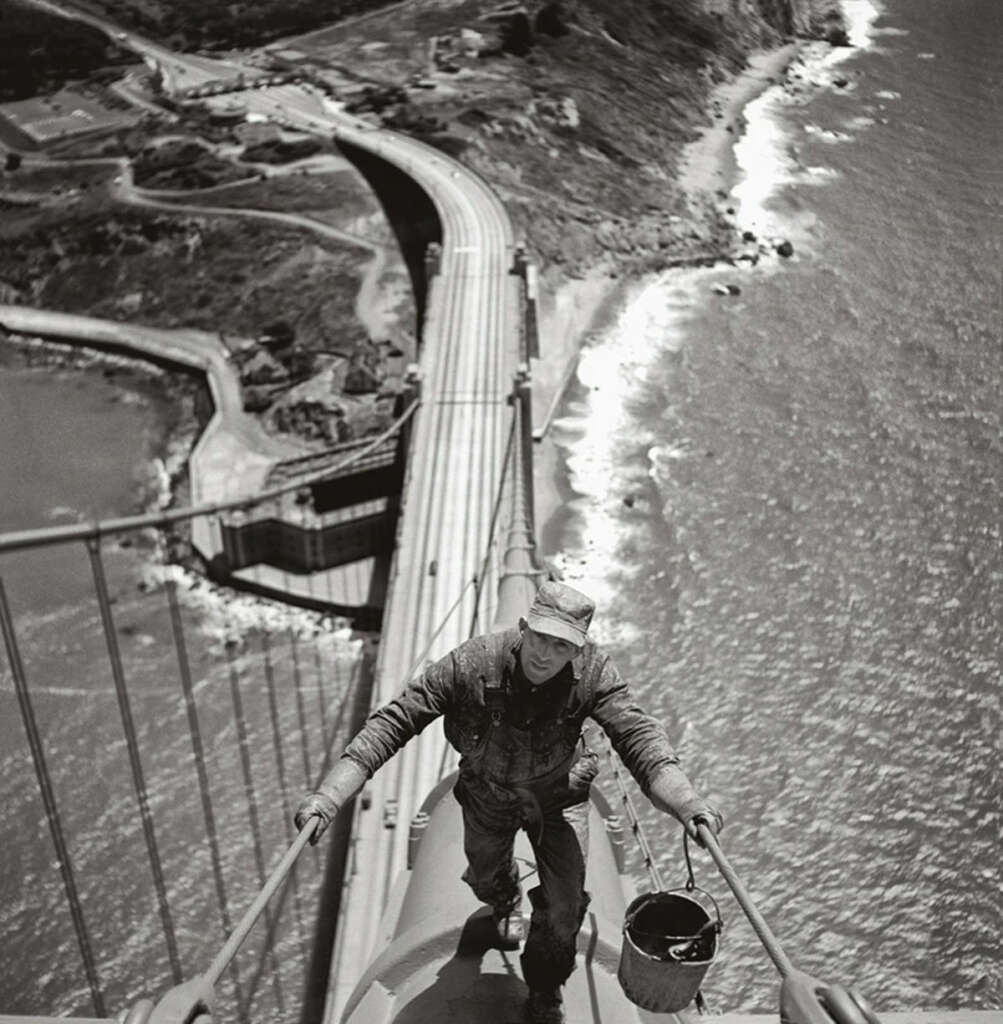 A construction worker stands on a high cable of the Golden Gate Bridge, holding a bucket in one hand. The bridge and surrounding landscape, including the coastline and roads, are visible below. The image is in black and white.