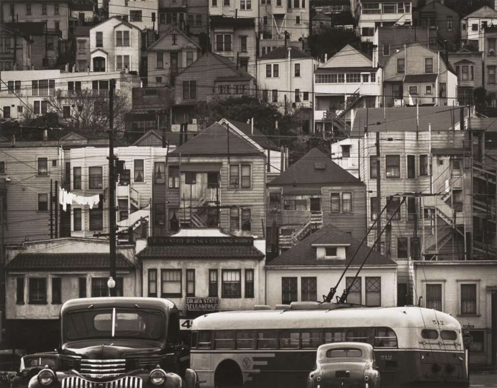 A black-and-white photo of a hillside densely packed with houses. In the foreground, a vintage bus and cars are parked on the street. Laundry hangs on a line between buildings, and signs of urban life are evident.