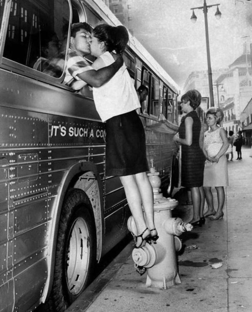 A woman stands on a fire hydrant to kiss a man through a bus window. The bus is parked on a city street with "IT'S SUCH A CO..." visible. Two other women and a child are waiting on the sidewalk nearby. The scene captures a moment of farewell or greeting.