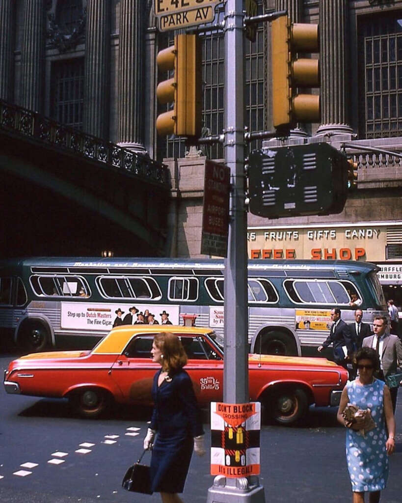 A vintage urban street scene showing a red car and a green bus with an advertisement passing by. People walk on the sidewalk in front of a building with signs for a gift and coffee shop. A pole with traffic lights and signs stands in the foreground.