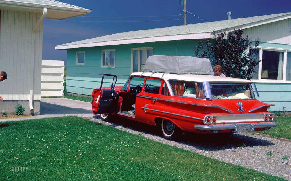 A vintage red station wagon is parked in a driveway beside a light green house. The car's doors are open, and it has a rooftop cargo bag. A person is partially visible near the car. The sky is clear and blue.