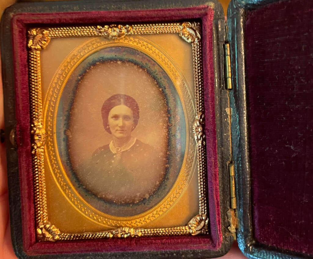 A vintage, sepia-toned photograph of a woman in an ornate, oval gold frame with intricate detailing. The frame is set in a velvet-lined, open case, with a deep red color on the right side. The woman is wearing a dark dress and a necklace.