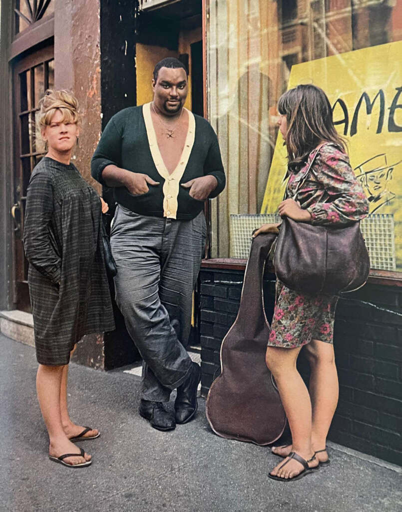 Three people stand on a city sidewalk near a storefront. The person in the middle leans against the building, wearing a cardigan and sandals. A woman in a patterned dress holds a guitar case, while another in a loose dress looks towards the camera.