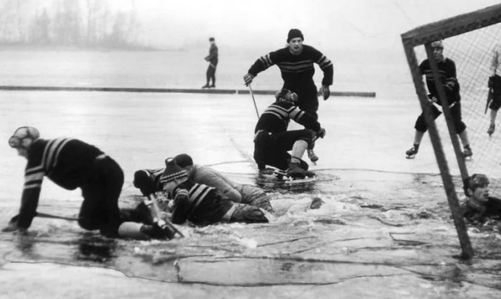 A black-and-white photo of people playing hockey on a frozen lake. Several players are scrambling near a cracked ice surface, some with skates on. A goal net is visible on the right, and a person in the background stands on the ice.