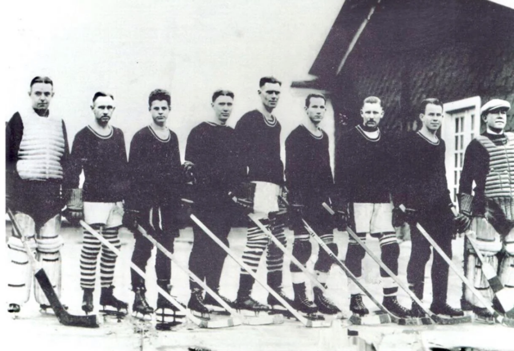 A vintage black-and-white photo of an ice hockey team. Nine players stand side by side outside, wearing matching dark sweaters and striped socks, holding hockey sticks. A building is partially visible in the background.