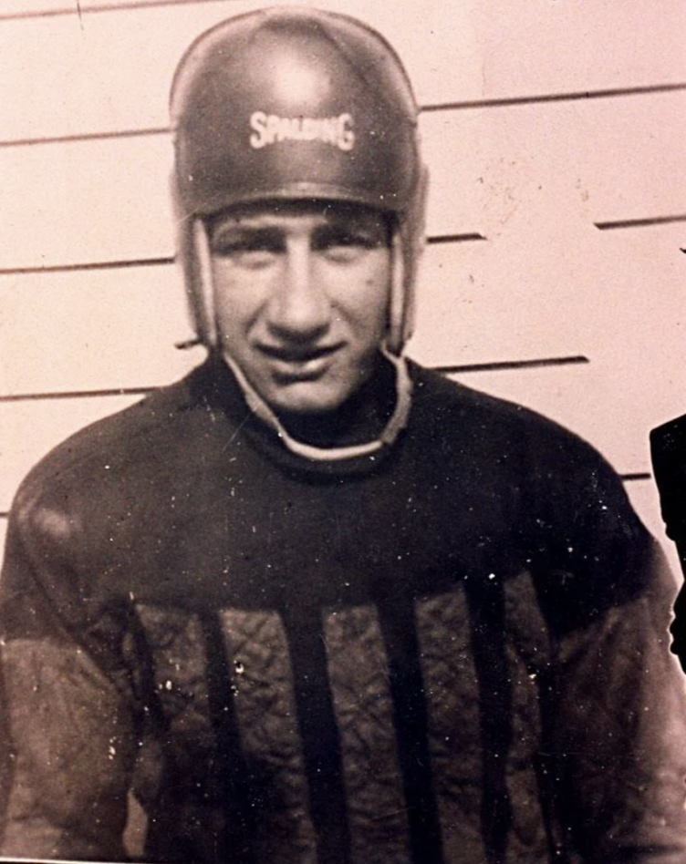 A vintage photo of a man wearing an old-style American football uniform, including a soft leather helmet with "Spalding" written on it. The background has horizontal lines resembling siding. The image is in black and white.