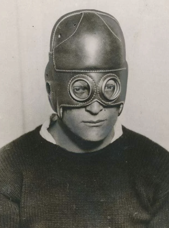 A person wearing a vintage leather aviator helmet with large round goggles. The helmet covers their head and ears, leaving only the face visible. They are dressed in a dark sweater, looking straight ahead with a neutral expression.