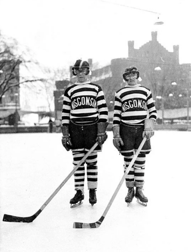 Two ice hockey players wearing striped jerseys labeled "Wisconsin" stand on an outdoor rink. They have skates, old-fashioned helmets, and hold hockey sticks. Snow is on the ground, with a large building visible in the background.