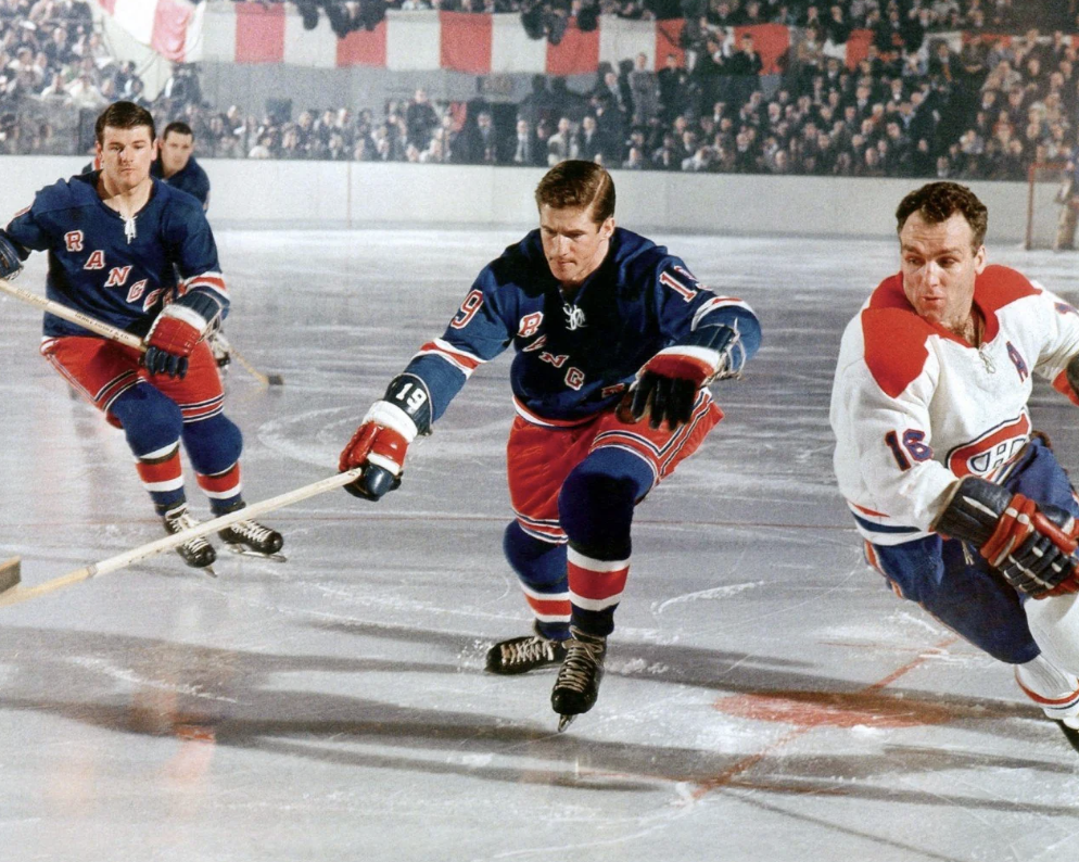 Three hockey players in action on the ice rink, wearing blue and red uniforms. One player in a white and red uniform skates beside them. A crowd of spectators watches from the stands in the background.