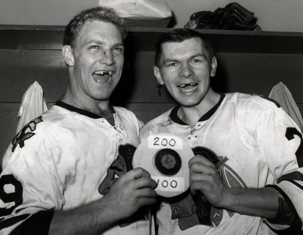 Two hockey players in jerseys smile while holding a celebratory puck inscribed with "200" and "100," in a locker room setting.