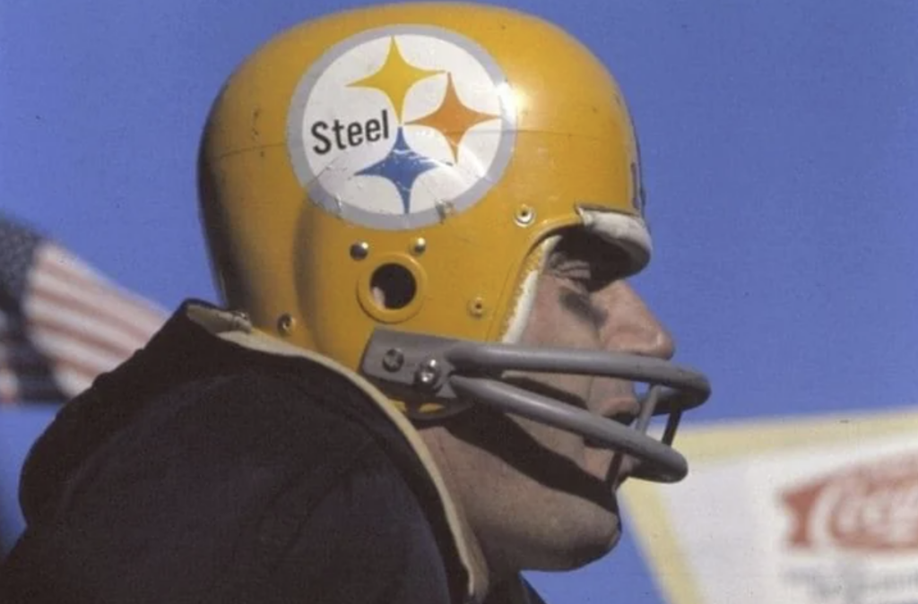 A football player in a yellow helmet with a Steel logo is captured in profile. The background features a clear blue sky, an American flag, and blurred signage.