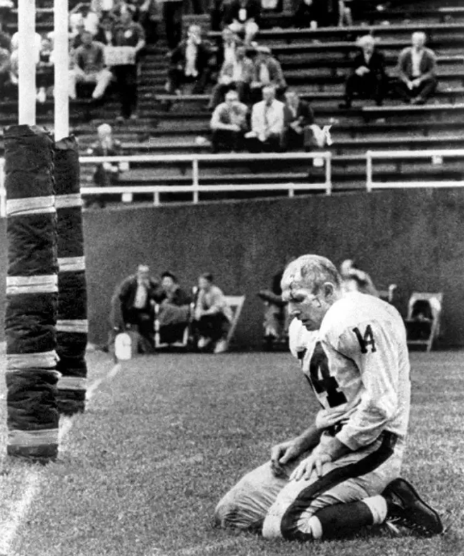 A football player in a number 14 jersey kneels on the field near the goalpost, helmet on, head down. The stands in the background have a few spectators. The image is black and white, conveying a sense of historical sports moment.