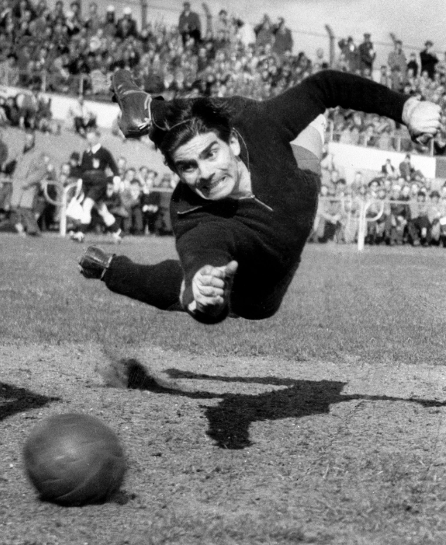 A goalkeeper in a vintage football match dives mid-air to stop a ball near the goal line. The crowd in the background watches intently, and the scene captures dynamic motion and determination. The image is in black and white.