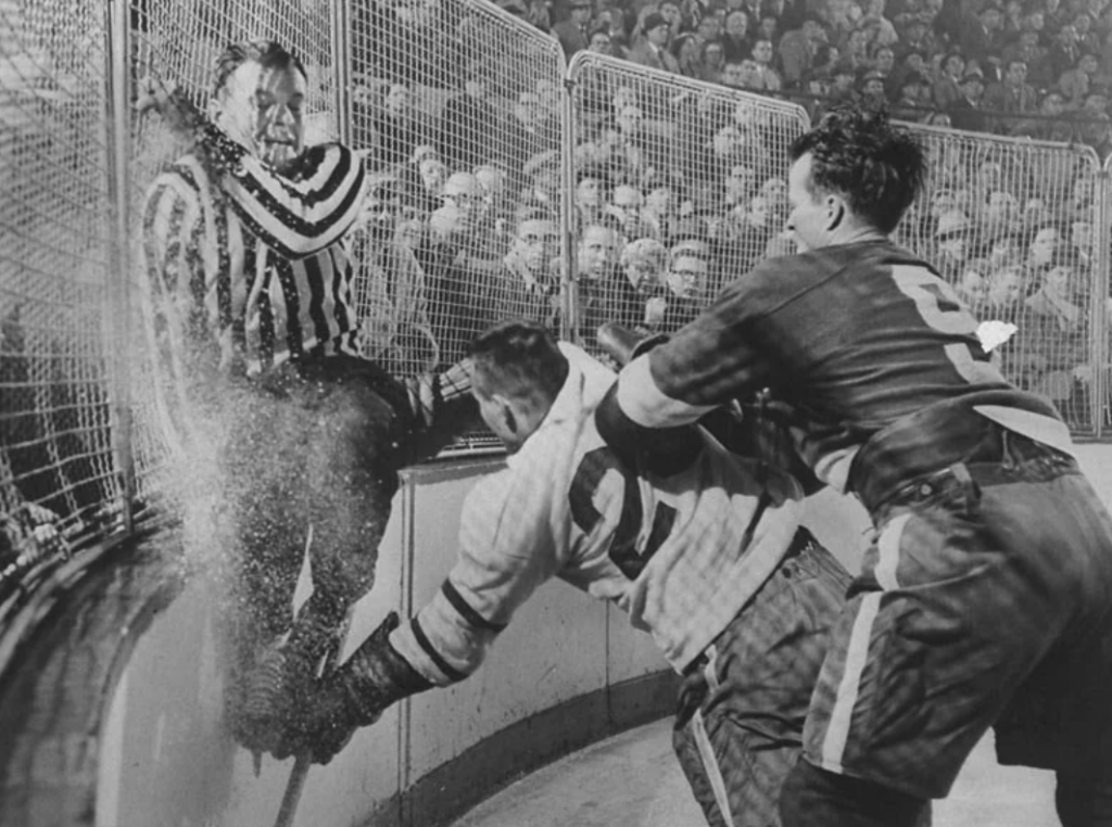 A black-and-white photo shows a hockey referee pressed against the rink fence as two players clash in front of a large crowd. One player has his arm around the other, who has fallen back slightly. The audience watches intently.