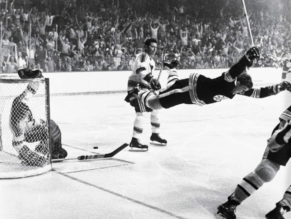 A hockey player in mid-air celebrates scoring a goal as the puck is in the net. The goalie sits dejectedly while other players watch. The background shows cheering fans in a packed arena. The scene captures the excitement of the moment.