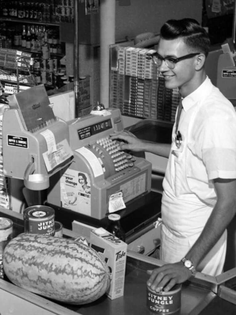 A young man wearing glasses operates a vintage cash register in a grocery store. A large watermelon, a can labeled "Jitney Jungle Coffee," and a box labeled "Tide" are on the counter. Shelves of products are visible in the background.