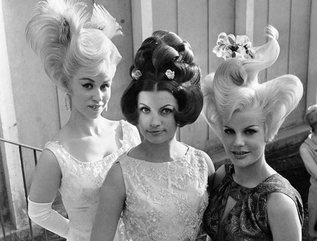 Three women in 1960s-style dresses showcase elaborate, gravity-defying hairstyles. The hairdos are sculpted with waves and curls, adorned with decorative elements. They pose confidently in front of a textured wall, embodying vintage fashion.