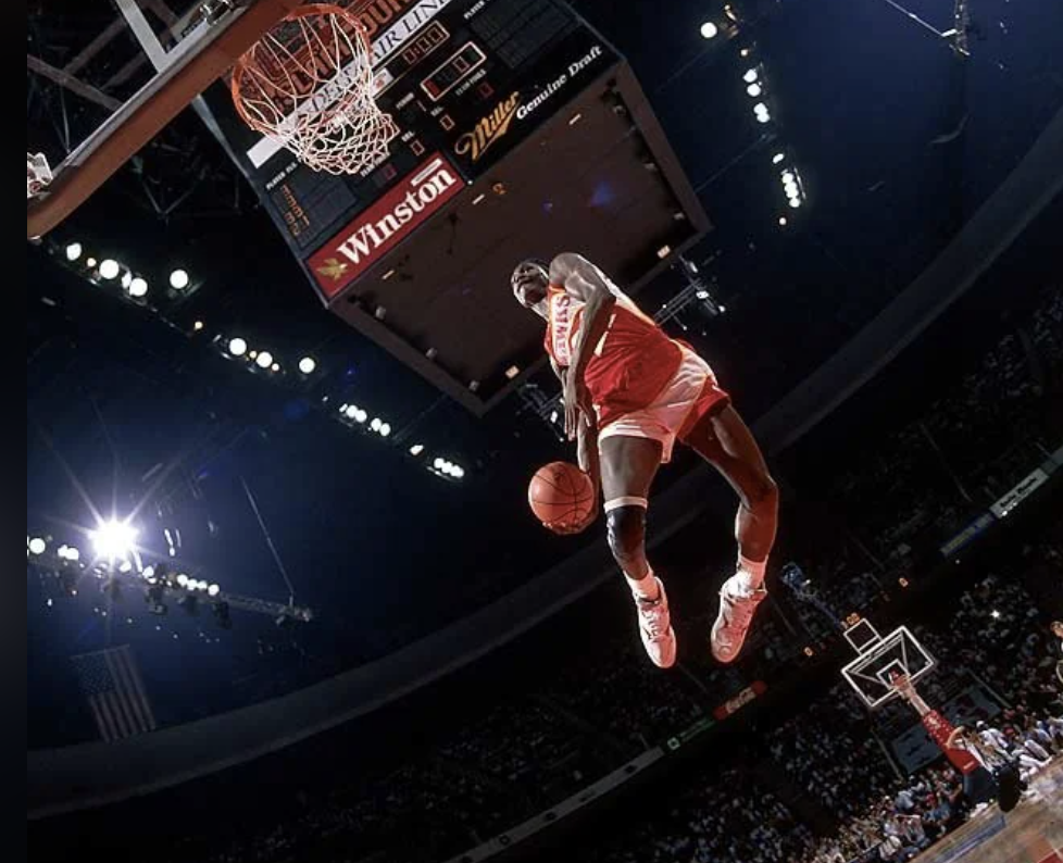 Basketball player in a red jersey performing a mid-air dunk, with the crowd and arena lights visible in the background.