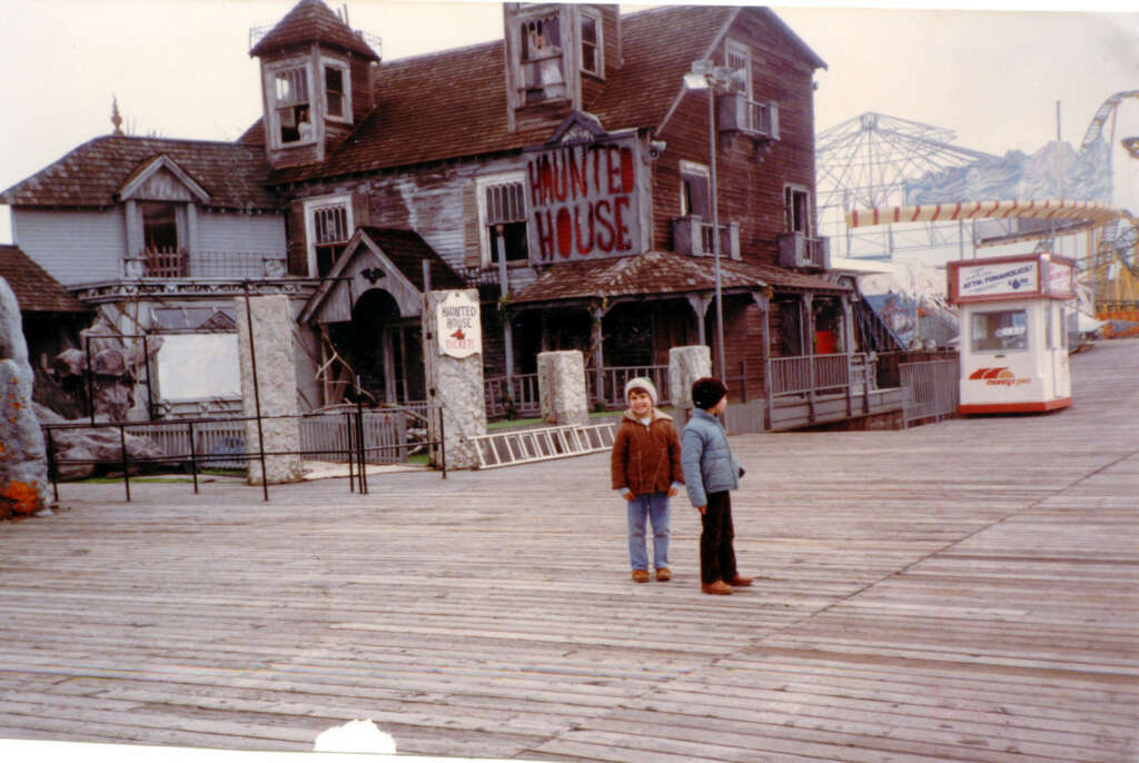 Two children stand on a wooden boardwalk near a decrepit haunted house attraction. The building is weathered with a sign that reads "Haunted House." An empty amusement park ride is in the background.