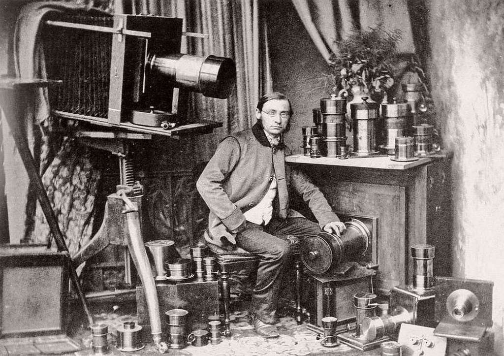 A man from the 19th century sits beside an early large-format camera with a tripod. He is surrounded by various camera lenses on a table and the floor. The setting has ornate curtains and a patterned rug.