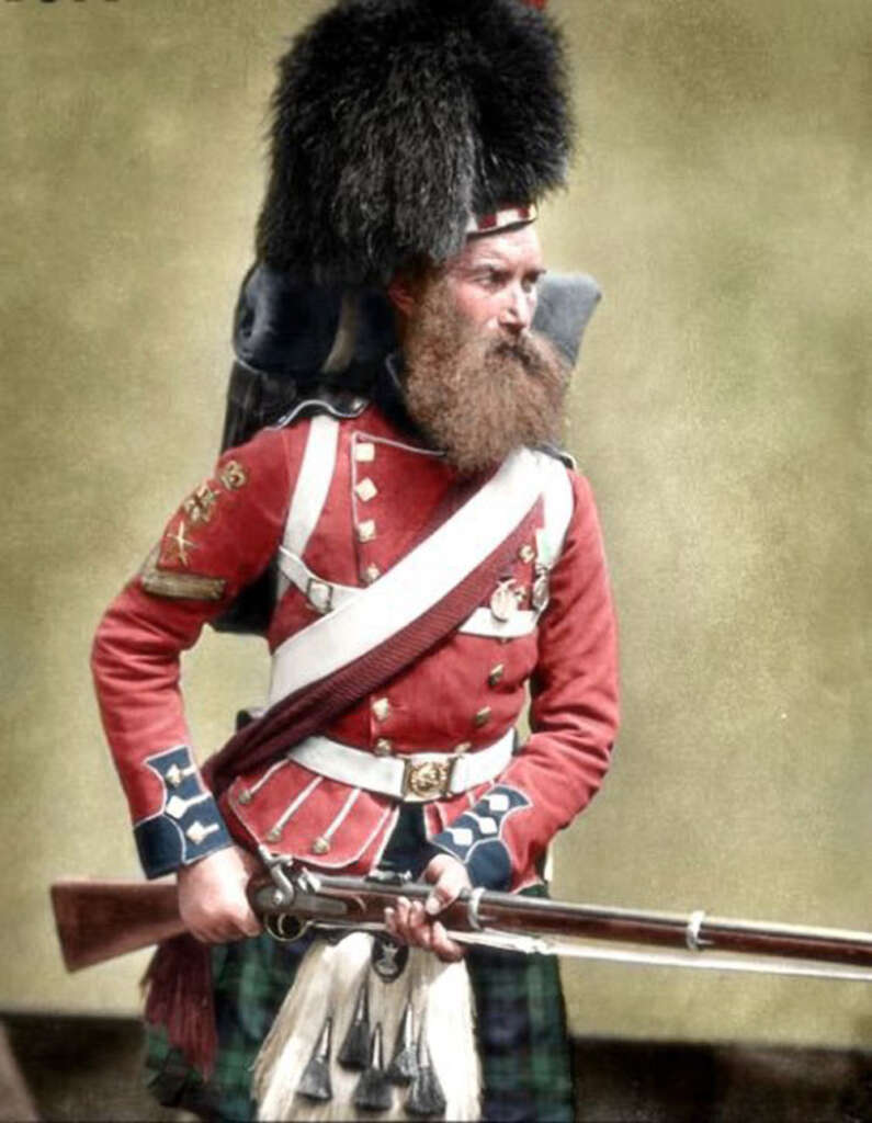 A historical image of a soldier in 19th-century British military attire. He wears a red coat with gold buttons, a black fur hat, and a kilt. The soldier holds a rifle and has a long beard, set against a plain background.