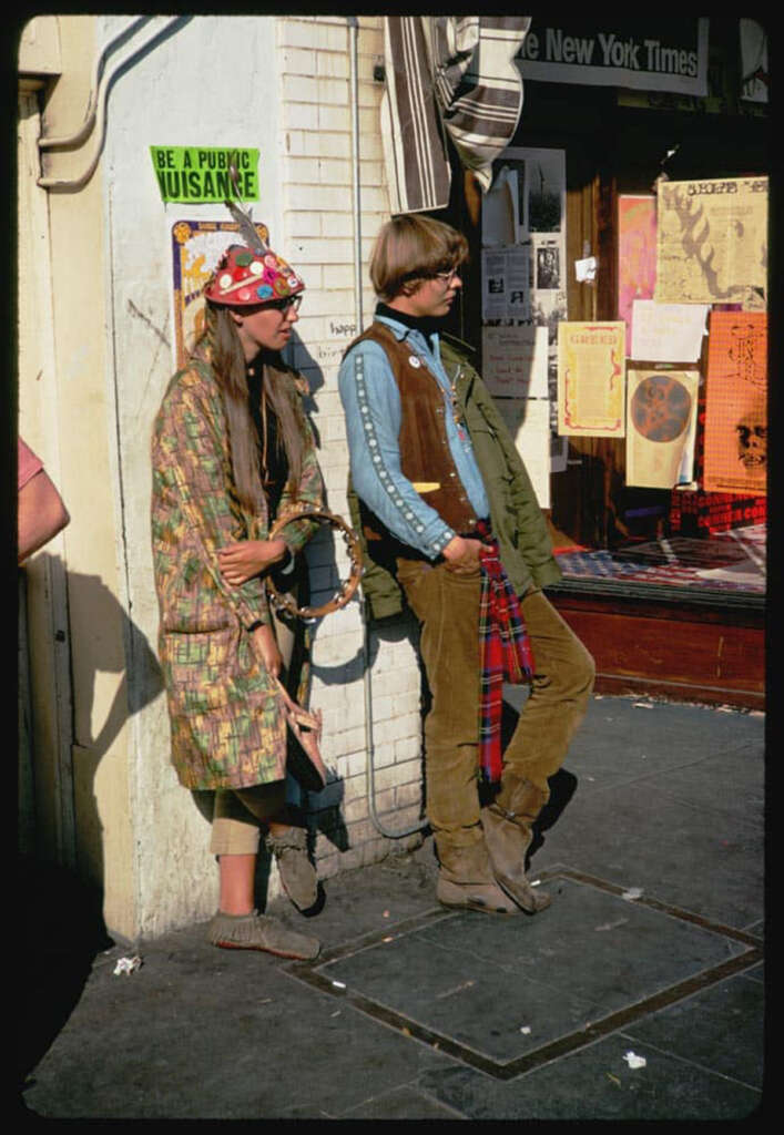 Two people in 1960s-style clothing stand on a street corner. One wears a patterned hat and coat, the other a denim jacket over a green shirt. Posters and signs cover the wall behind them. A sign reads "BE A PUBLIC NUISANCE.