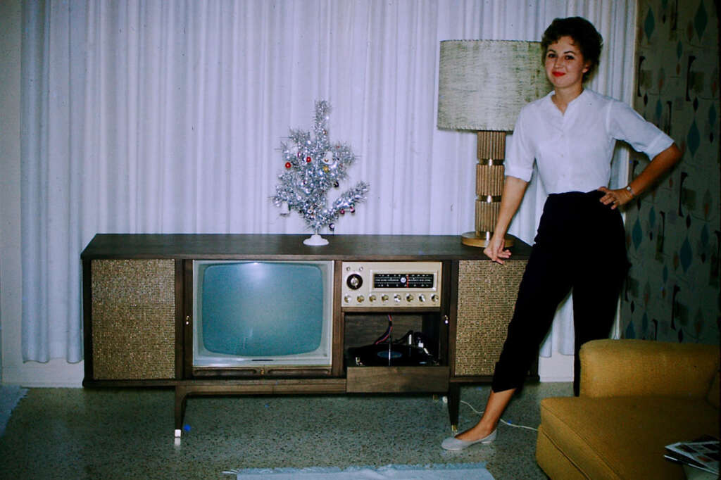 A woman stands beside a vintage television and radio console in a living room. She is wearing a white blouse and black pants. A small silver Christmas tree and a lamp are on the console. White drapes and a beige couch are in the background.