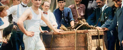 A group of men surrounds a wooden crate outdoors. A man in the foreground, wearing a white sleeveless undershirt and shorts, confidently stands next to the crate. Others are dressed in early 20th-century attire, including hats and suspenders.