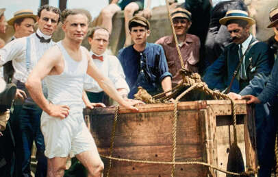 A group of men surrounds a wooden crate outdoors. A man in the foreground, wearing a white sleeveless undershirt and shorts, confidently stands next to the crate. Others are dressed in early 20th-century attire, including hats and suspenders.