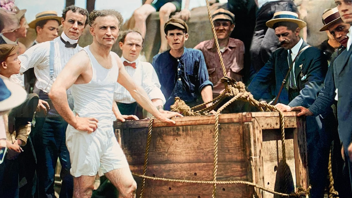 A group of men surrounds a wooden crate outdoors. A man in the foreground, wearing a white sleeveless undershirt and shorts, confidently stands next to the crate. Others are dressed in early 20th-century attire, including hats and suspenders.