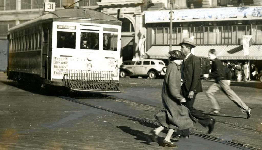 A vintage street scene featuring a streetcar labeled "31" approaching as two people walk across the tracks. They're dressed in early 20th-century attire. A man runs in the background, with buildings and old cars visible.