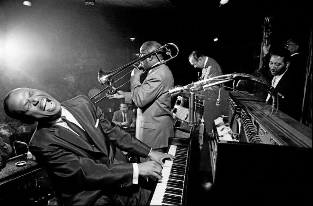 A black-and-white photo of a jazz band performing. The pianist, playing energetically, is leaning back with a joyful expression. A trombonist, saxophonist, and bassist are also engaged in the performance, creating an energetic atmosphere.